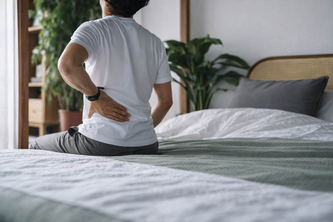 Man sitting on the edge of a bed holding his lower back in discomfort